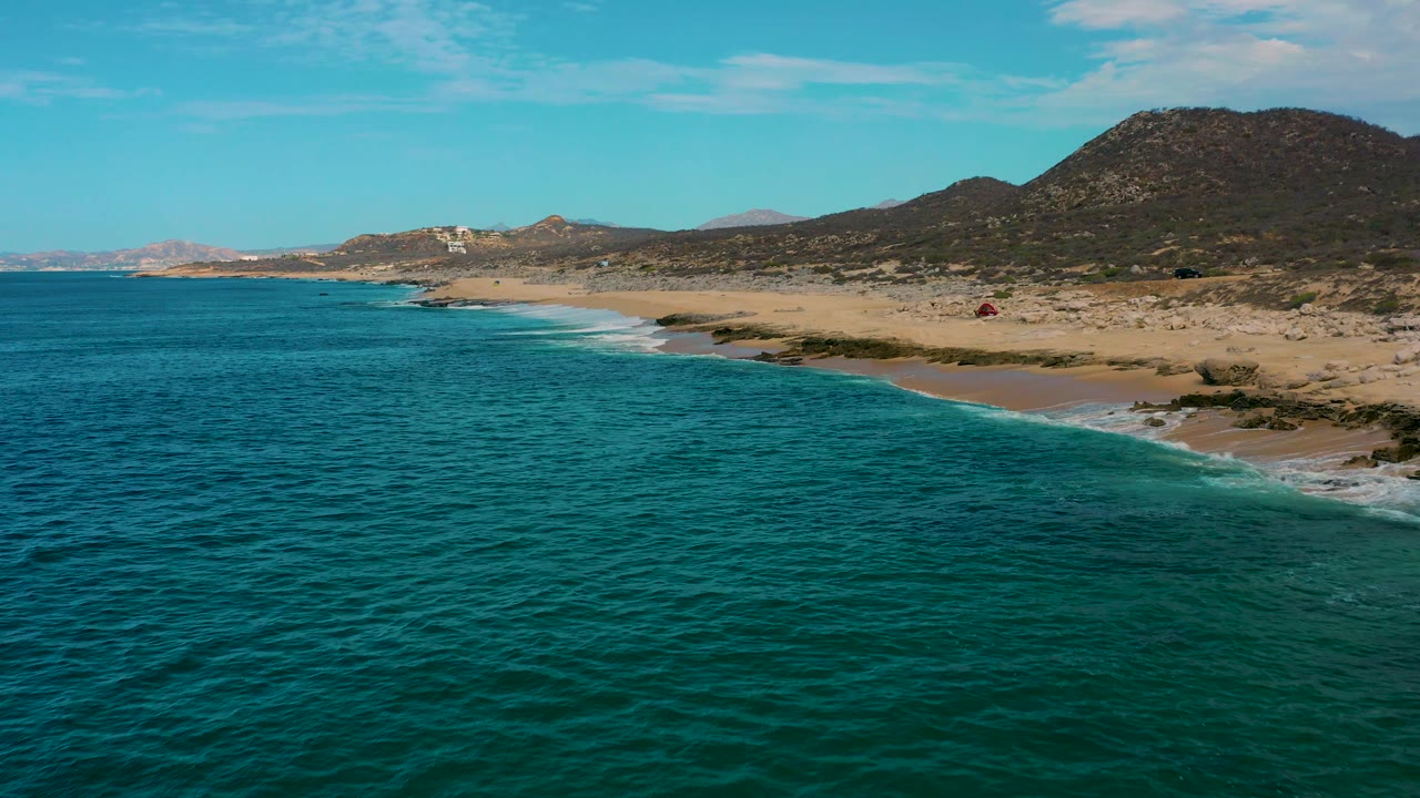 Aerial frame at 0:02 over Laguna Beach, Baja California Sur, with a wide band of turquoise water, pale sand curving along the shore, dark hills to the right, and no visible people.
