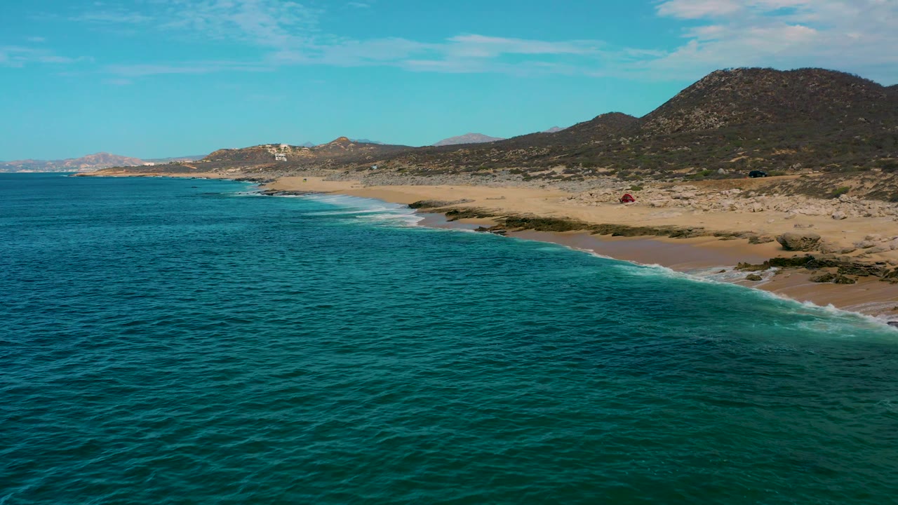Aerial frame at 0:03 over Laguna Beach, Baja California Sur, where the ocean foreground leads into a foamy shore break, sandy beach, scrub, rocky hillside, and no visible people.