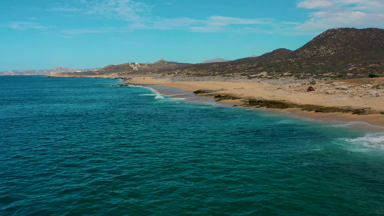 Aerial frame over Laguna Beach, Baja California Sur, showing turquoise-green water, a pale sand beach, distant rocky coast, scrubby land, dark hills, and no visible people.