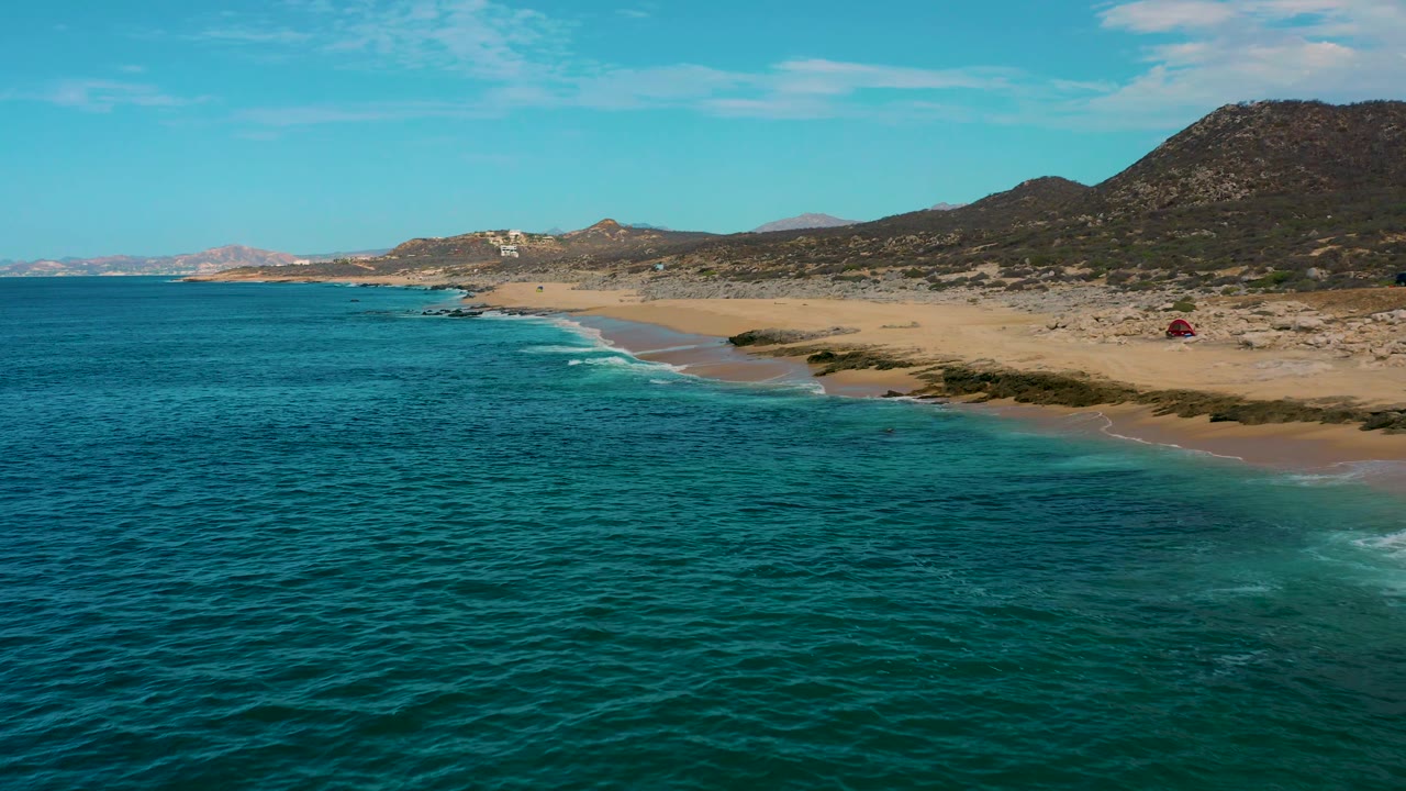 Laguna Beach aerial still with textured green water across the lower frame, a thin white surf edge along the beach, scrub-covered sand, hills to the right, and no visible people.