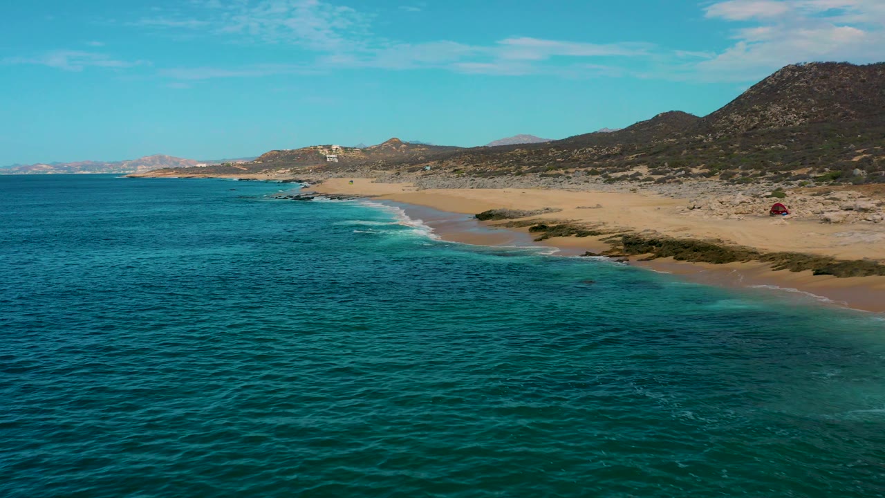 Drone-height view of Laguna Beach in Baja California Sur with dark green water in front, mild surf on the sand, open coastal scrub, a hillside backdrop, and no visible people.