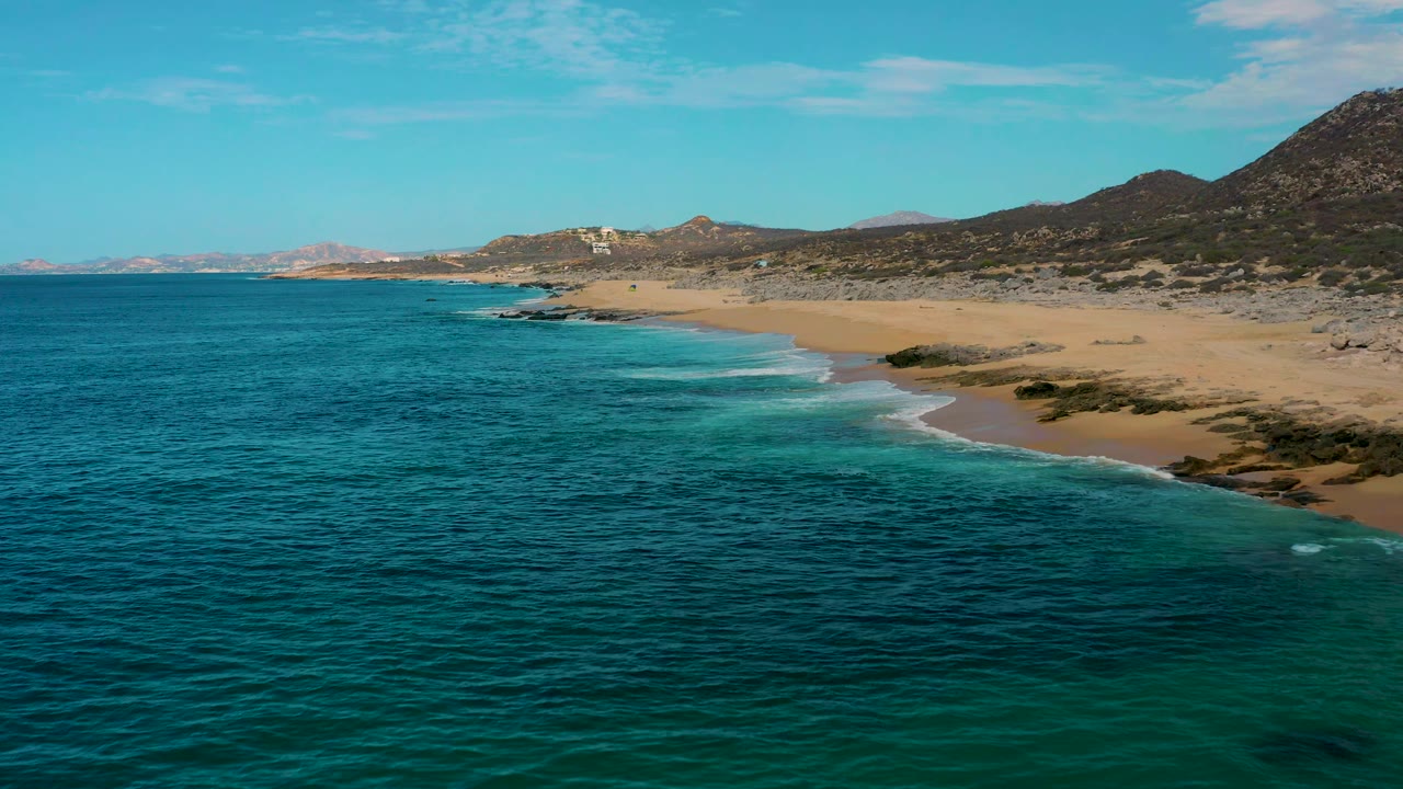 Drone view along Laguna Beach with turquoise shallows spreading near the sand, darker ocean at left, rocky hills inland, and no visible people.