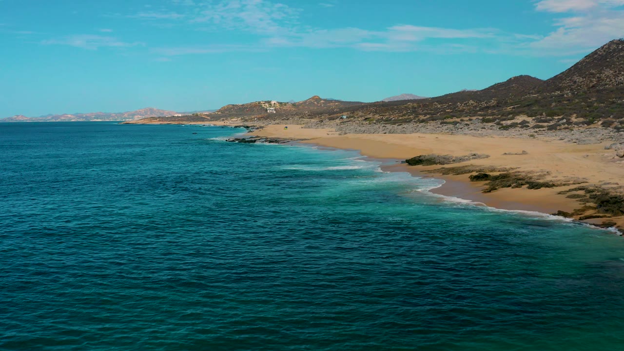 Wide aerial frame at Laguna Beach, Baja California Sur, showing ocean across most of the view, surf curling onto sand at right, inland hills, and no visible people.