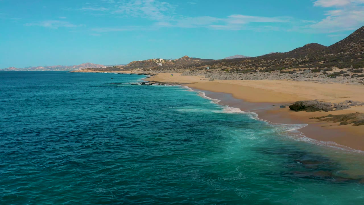 Aerial frame of Laguna Beach, Baja California Sur, where pale turquoise surf spreads toward a broad sandy beach beneath rocky coastal hills, with no visible people.
