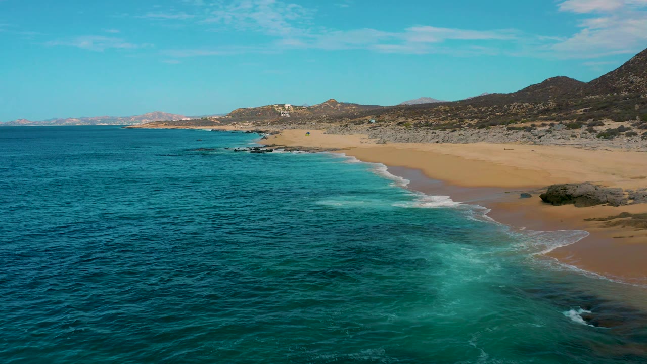 Aerial view of Laguna Beach, Baja California Sur, with darker ocean in the foreground, a bright patch of breaking surf near shore, tan sand at right, and no visible people.