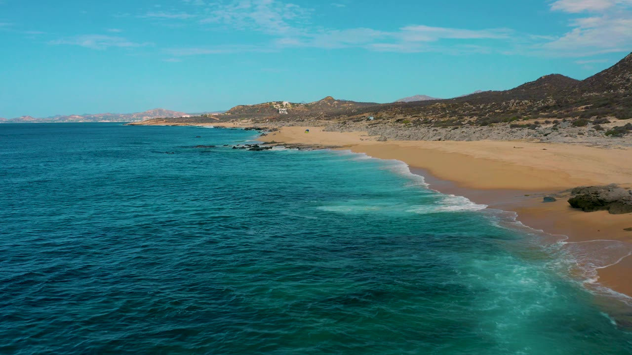 Aerial frame over Laguna Beach, Baja California Sur, showing a diagonal sandy shoreline, blue ocean to the left, small foamy waves, rocky terrain inland, and no visible people.