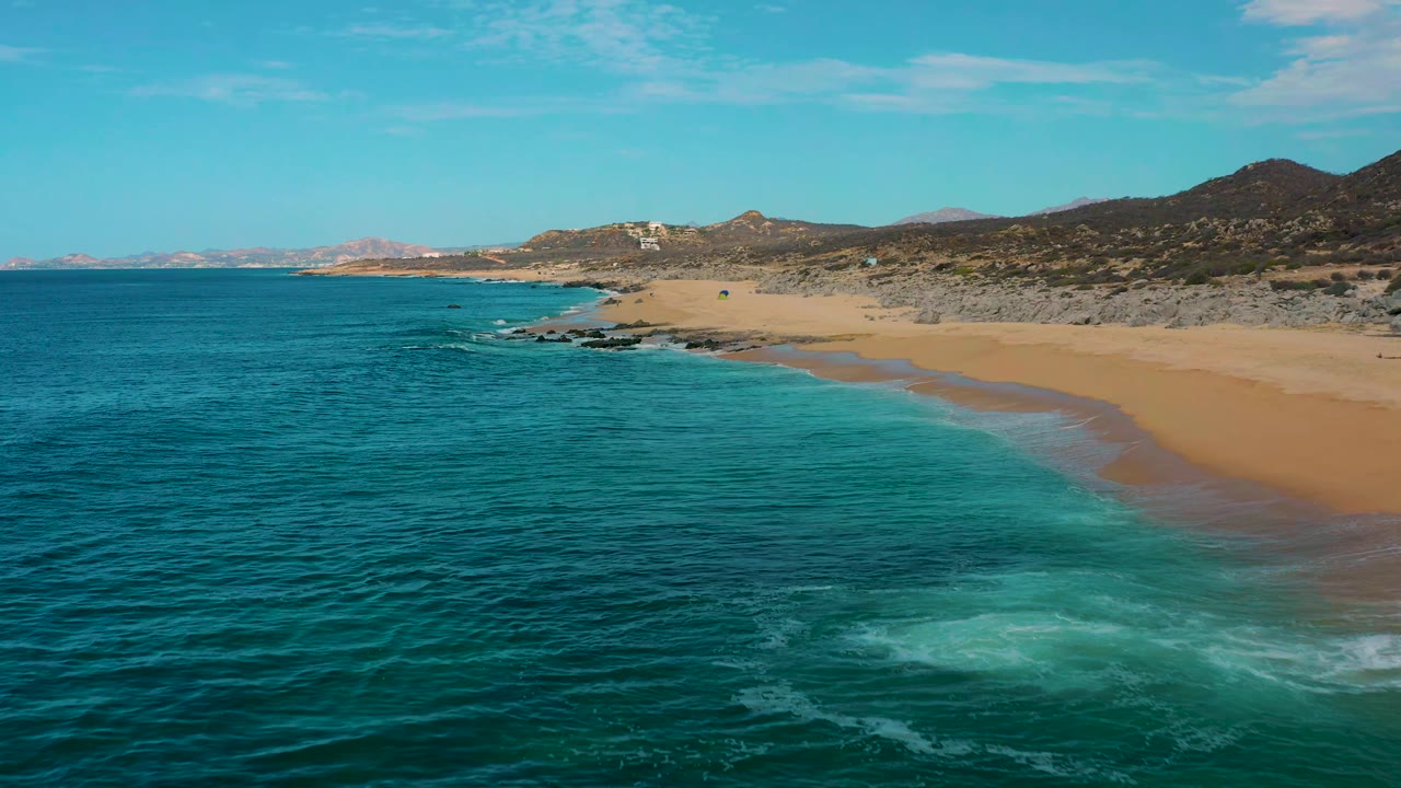 Aerial view at Laguna Beach, Baja California Sur, Mexico, looking along teal water toward a tan beach, with a pale surf patch in the foreground and no visible people.