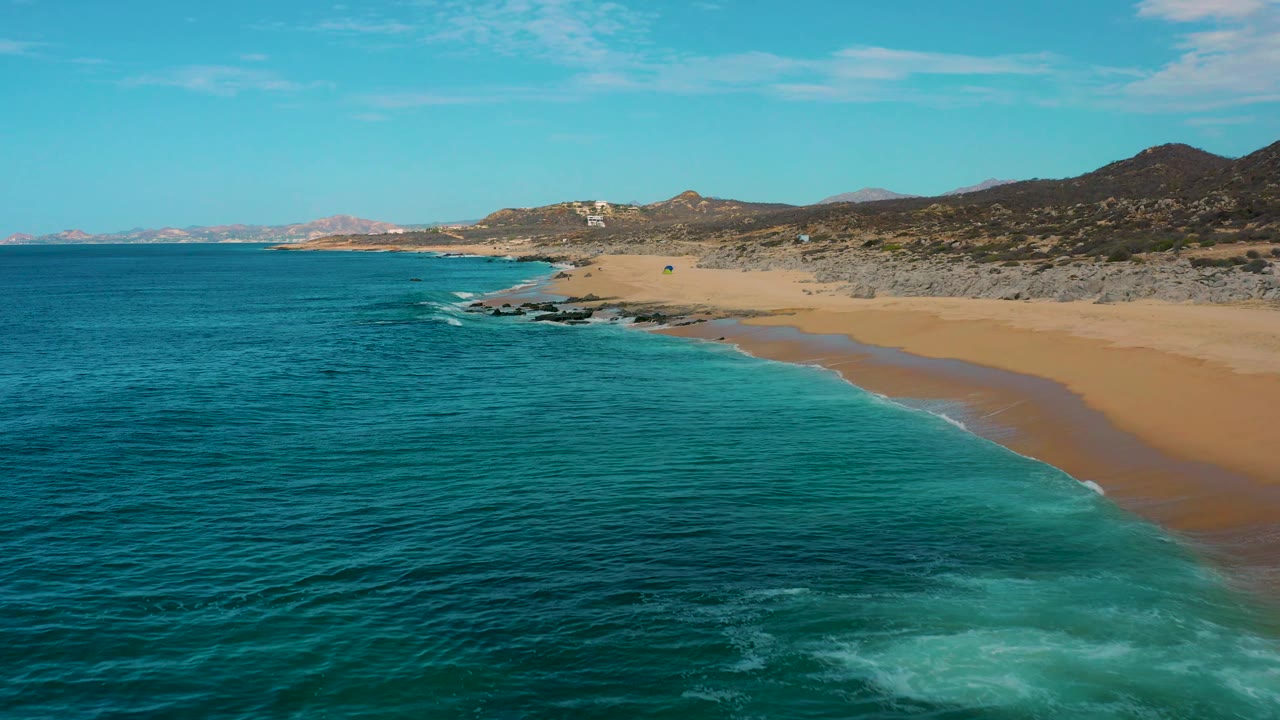 Aerial frame over Laguna Beach, Baja California Sur, Mexico, with darker teal ocean filling more of the foreground, a curving strip of surf on the right, rocky shoreline ahead, and no visible people.