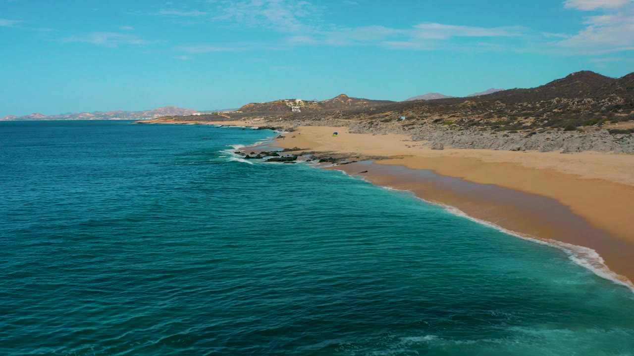 Aerial view at Laguna Beach, Baja California Sur, Mexico, looking down the coast as teal ocean fills the left side, a narrow white wave line traces the beach, and no visible people appear on the sand.