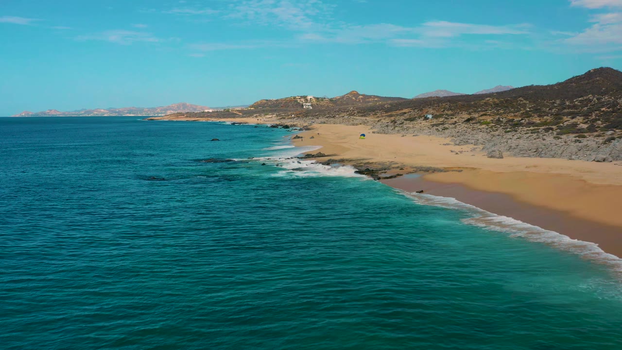 Aerial coastal frame from Laguna Beach, Baja California Sur, showing darker blue water at left, lighter turquoise shallows near shore, pale sand on the right, and no visible people.