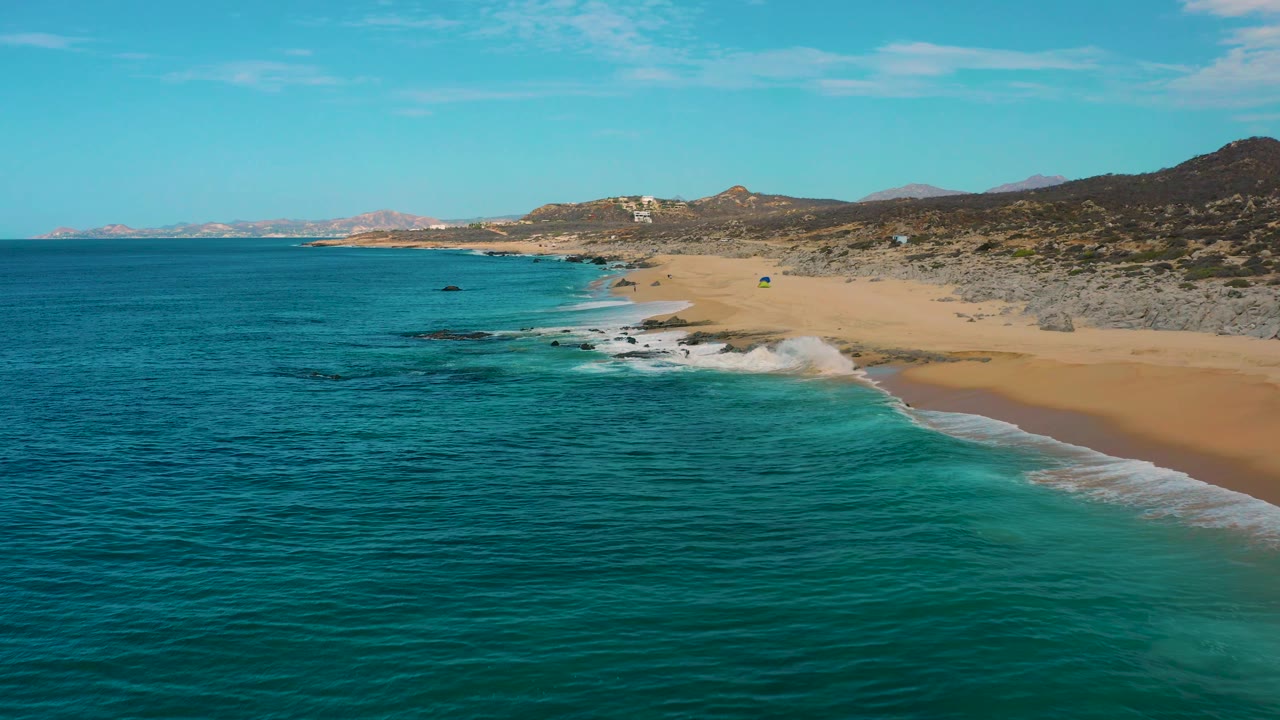 Drone view along Laguna Beach in Baja California Sur, Mexico, with white waves breaking near dark rocks, a long sand beach curving into the distance, arid hills behind it, and no visible people.