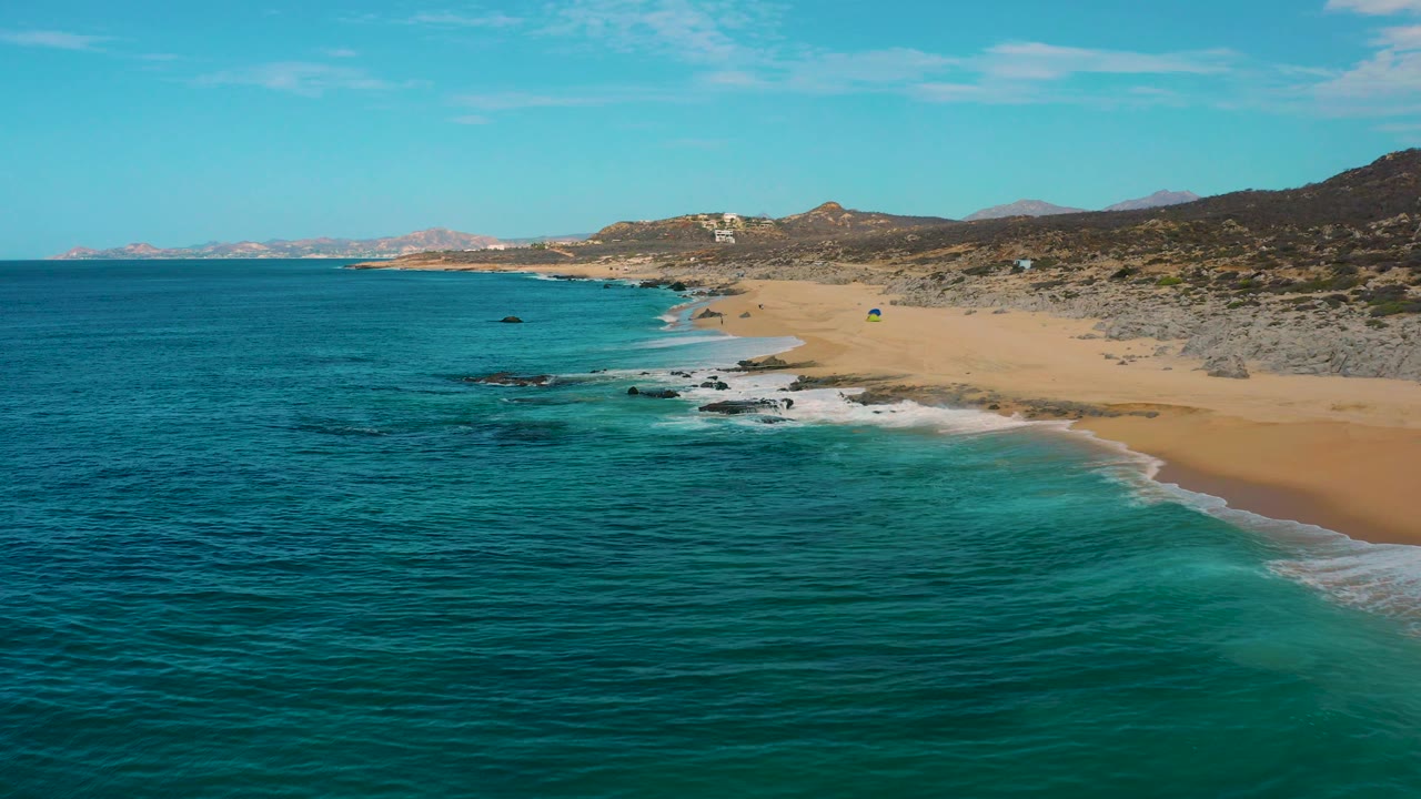 High aerial view of Laguna Beach, Baja California Sur, with rippled ocean filling the foreground, foamy surf at the beach edge, scattered rocks near shore, hillside terrain in back, and no visible people.