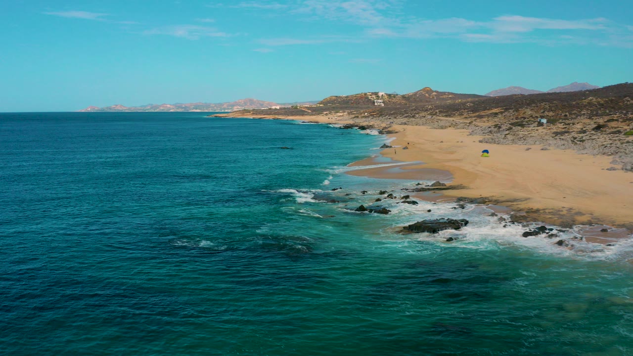 Aerial frame over Laguna Beach, Baja California Sur, Mexico, showing turquoise water, a sandy shoreline, dark rocks in the surf, low hills inland, and no visible people.