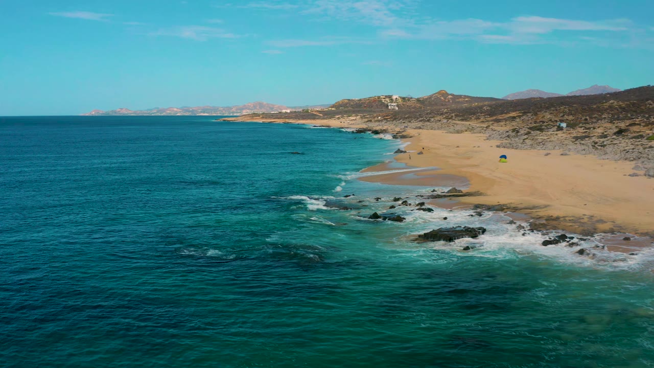 Drone view over Laguna Beach, Baja California Sur, Mexico, with blue-green ocean on the left, foamy waves over black rocks, tan beach on the right, and no visible people.
