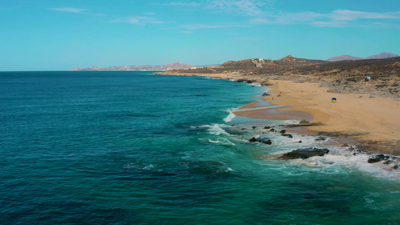 Aerial view at Laguna Beach in Baja California Sur, Mexico, showing a wider sweep of ocean, breaking waves along scattered rocks, a broad sandy beach, and no visible people.