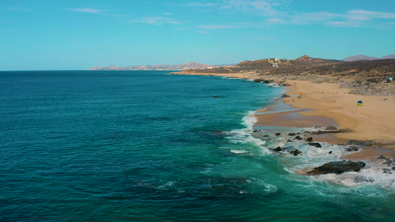 Laguna Beach, Baja California Sur, Mexico, from above, with teal water filling most of the frame, a tan beach curving along the right, rocky surf in the foreground, and no visible people.