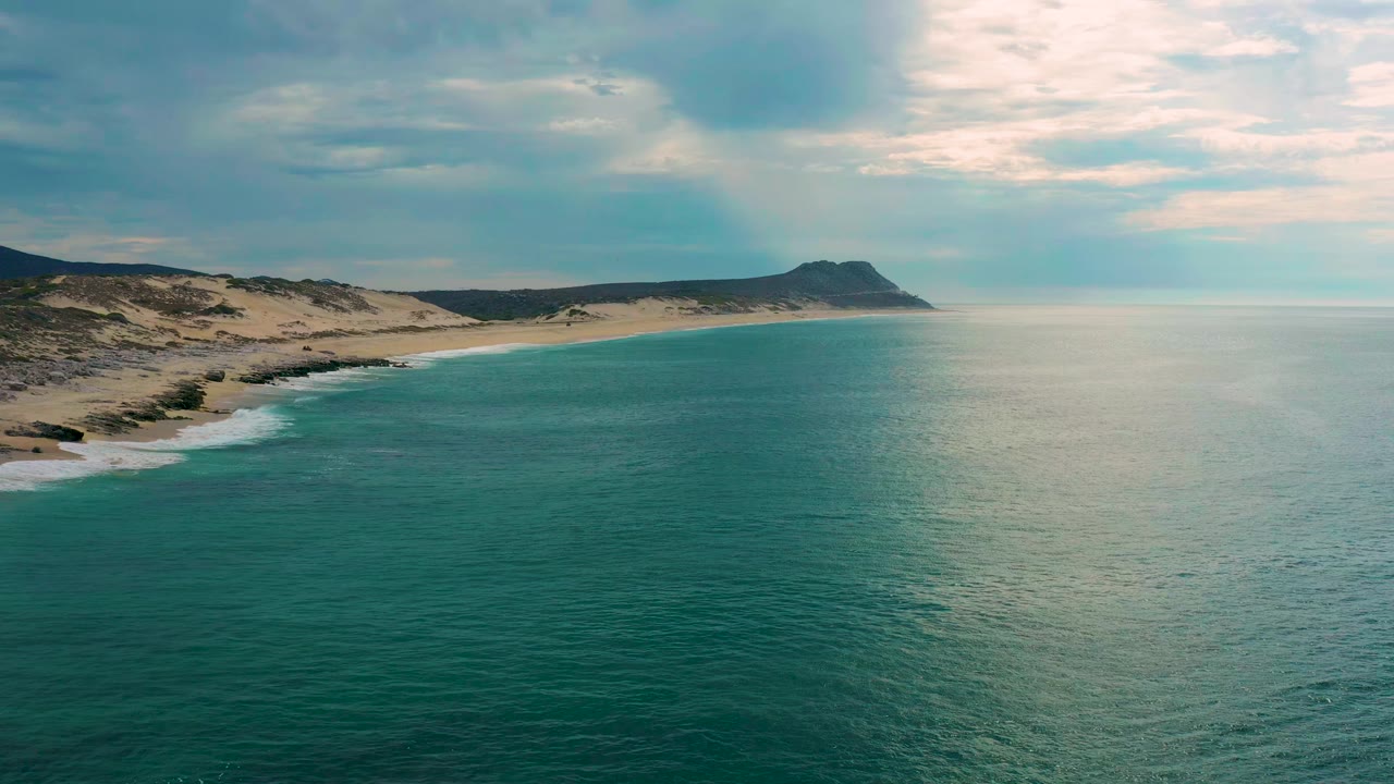 Wide aerial view at Laguna Beach, Baja California Sur, with teal sea in the foreground, sandy coast to the left, a distant headland, clouded sky, and no visible people.