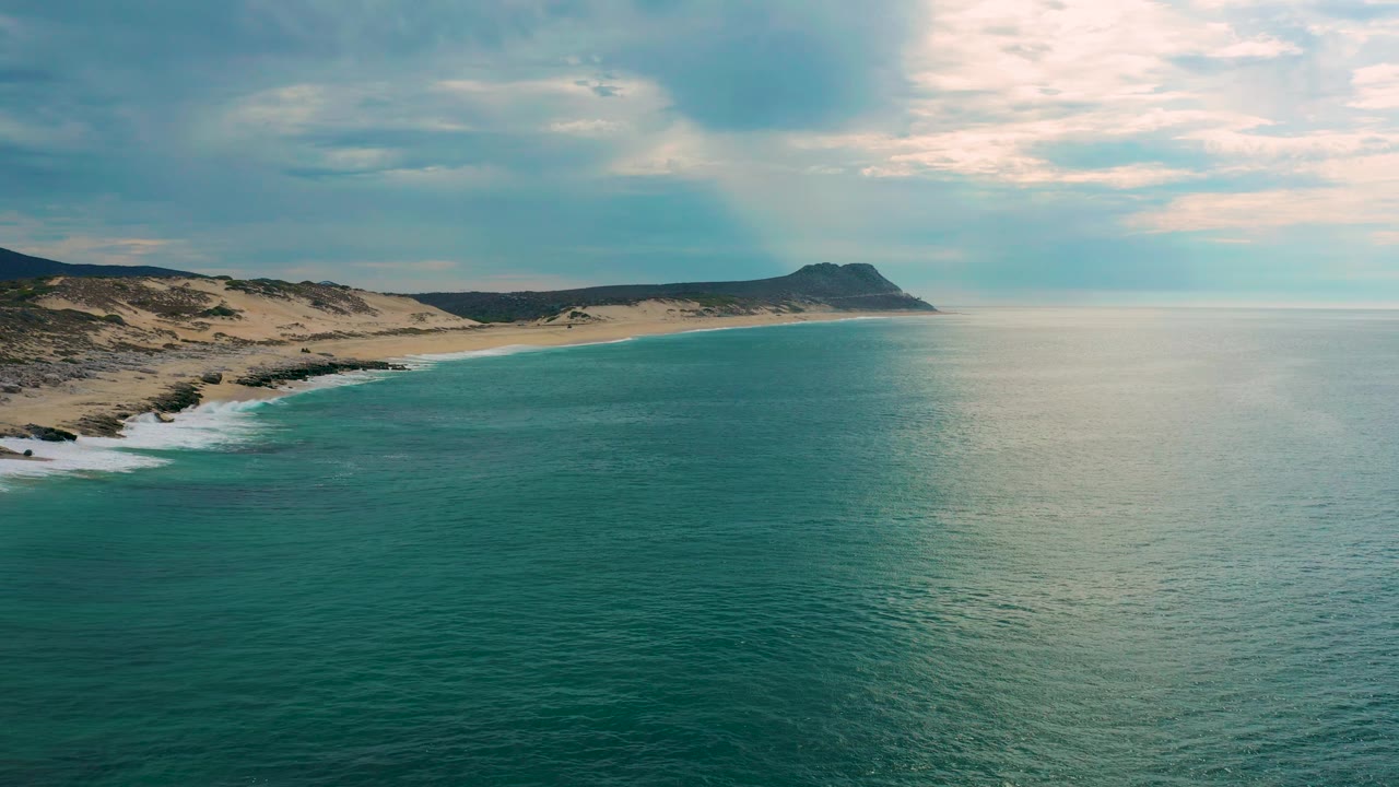 Mid-clip aerial frame at Laguna Beach, Baja California Sur, with pale beach and scrubby dunes to the left, teal water across the foreground, gray-white clouds above, and no visible people.