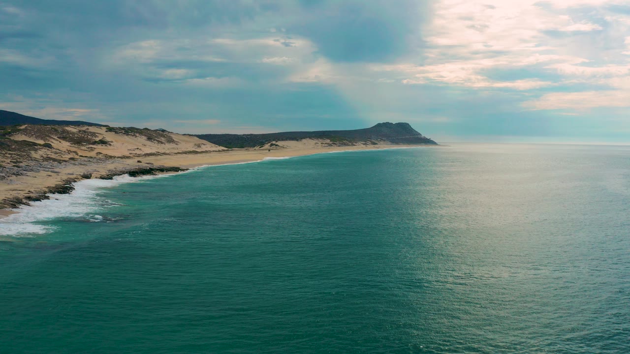 Aerial view over Laguna Beach, Baja California Sur, showing turquoise sea, pale surf on the left shoreline, sandy dunes, a distant headland, soft cloud cover, and no visible people.