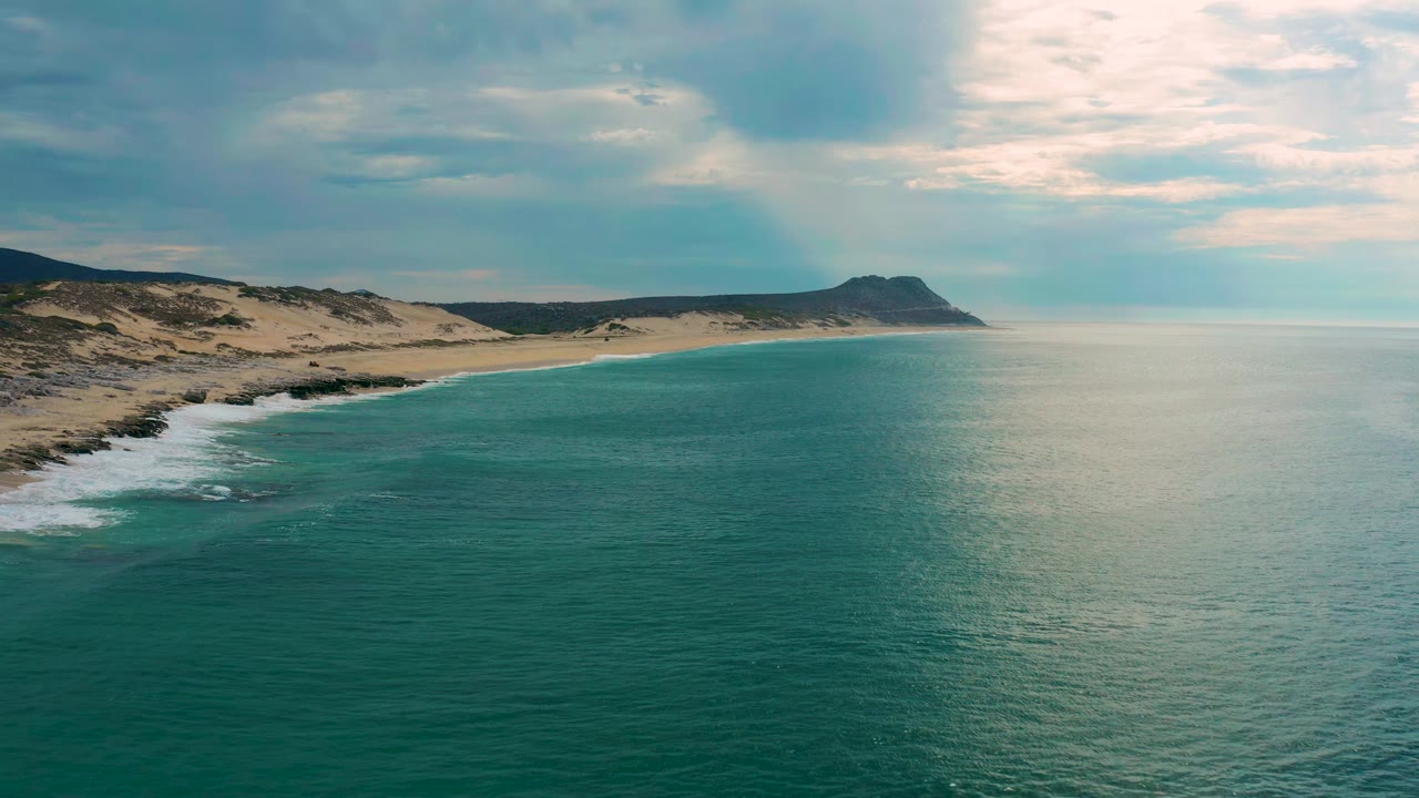 A second aerial frame over Laguna Beach, Baja California Sur, keeps the shoreline along the left edge, broad green-blue water across the foreground, a low headland in the distance, and no visible people.
