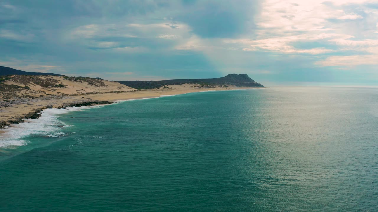 Aerial coastline at Laguna Beach, Baja California Sur, with small white breakers reaching the sandy edge, darker rocks near the shore, muted clouds overhead, and no visible people.