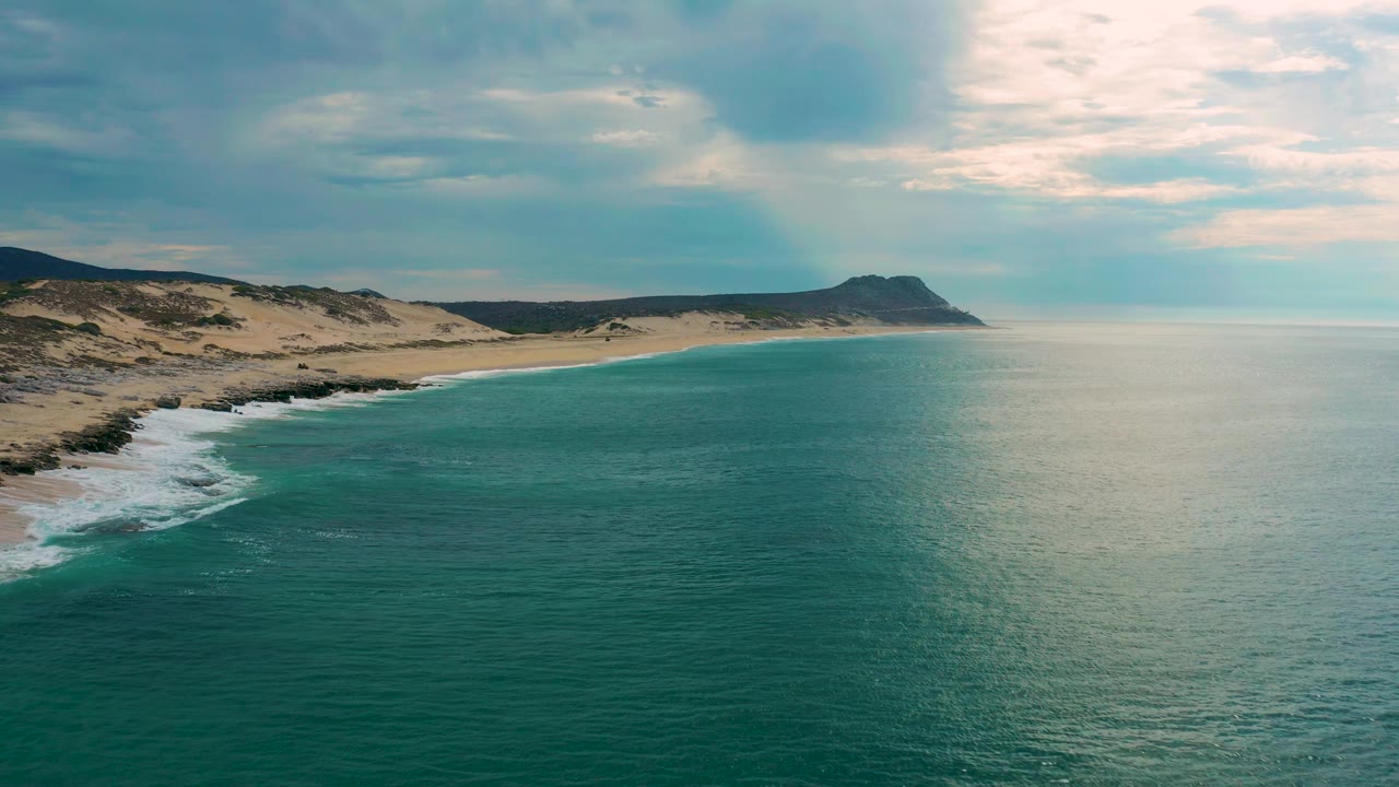 Mid-clip aerial view in Laguna Beach, Baja California Sur, showing the curving sandy coast on the left, broad calm water in front, pale sunlight near the horizon, and no visible people.