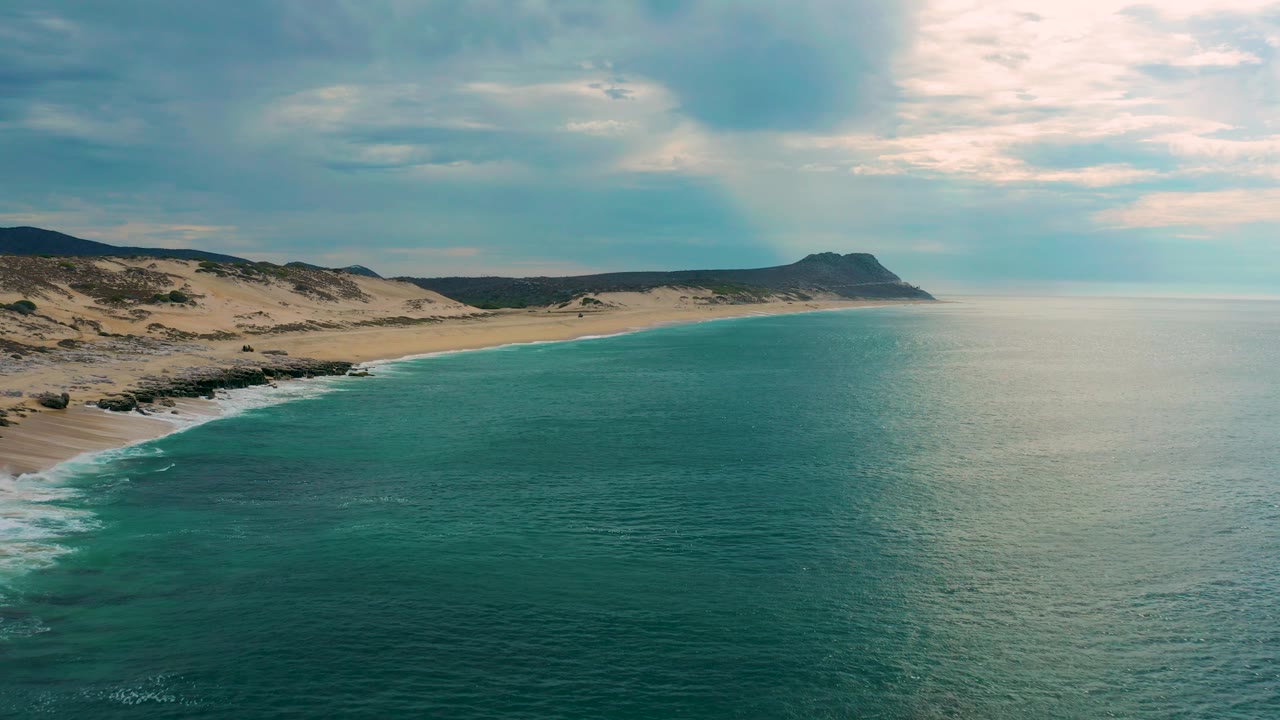 A wide aerial view of Laguna Beach, Baja California Sur, with more open water visible to the right, the sand and dunes tracing the left edge, and no visible people.