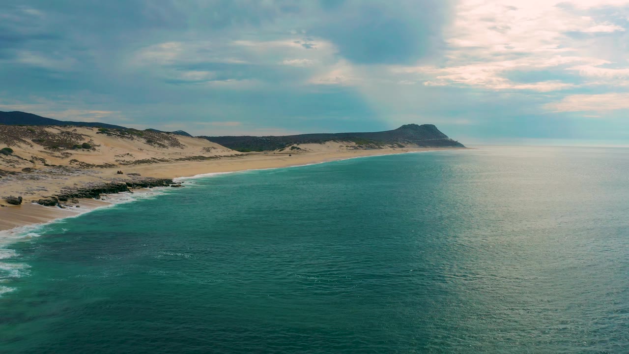 Ocean and sandy shore viewed from above under a bright cloudy sky.