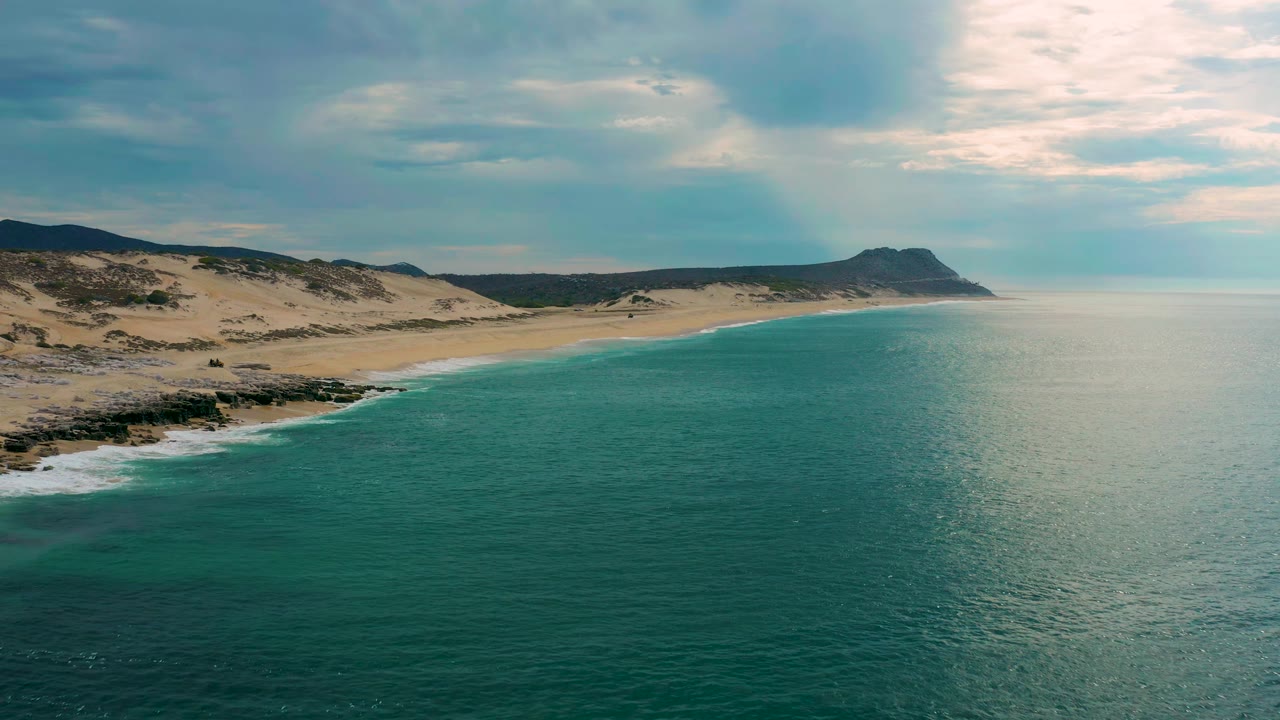Second aerial frame of Laguna Beach, Baja California Sur, holding a wide view of blue-green water, a curving sandy shore, dune ridges, dark rocks near the surf, and no visible people.