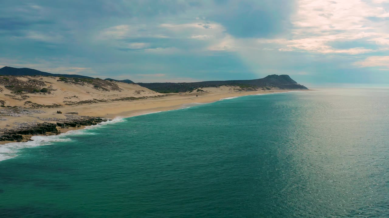 Mid-clip aerial view of Laguna Beach, Baja California Sur, showing dark rocks and white surf at lower left, sandy beach and dunes beyond, teal water across the frame, and no visible people.