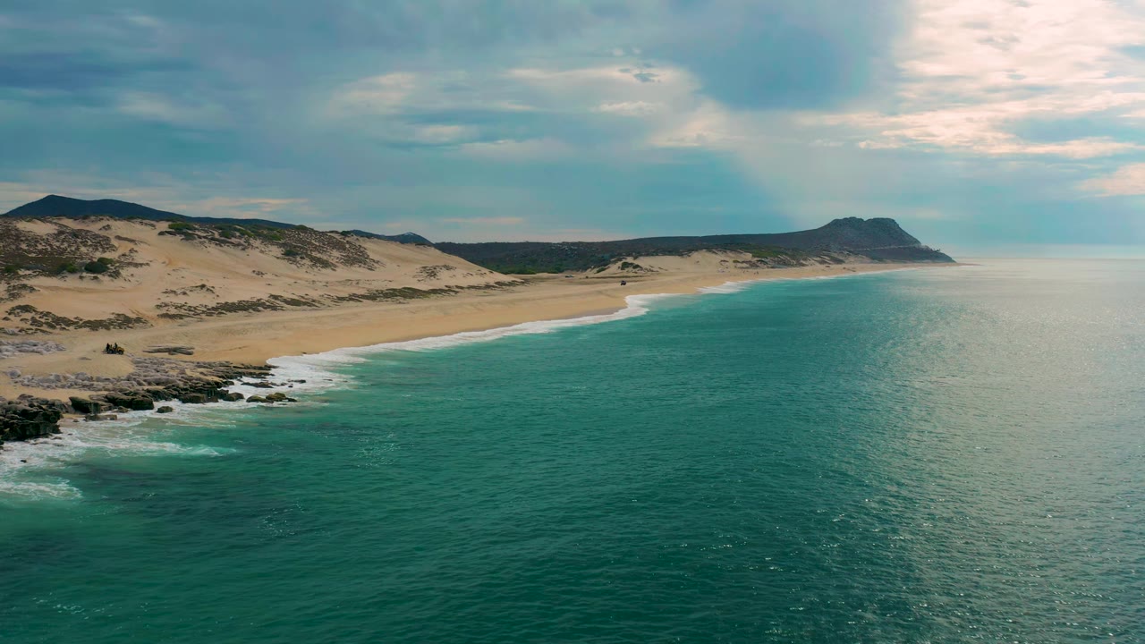 Laguna Beach coastline seen from above, with blue-green water filling the lower frame, a long sand beach curving toward a dark headland, and no visible people.