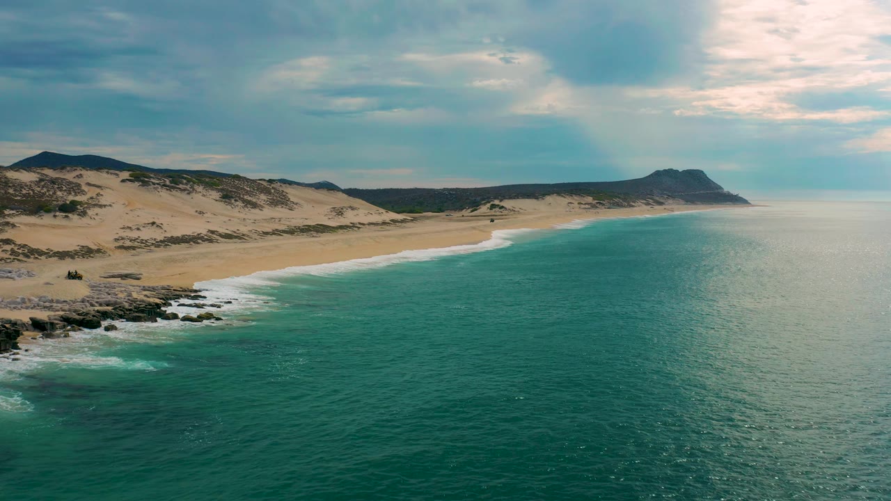 Drone-style aerial over Laguna Beach, Baja California Sur, where calm turquoise water meets a broad sand beach backed by dunes, low hills, and a distant headland with no visible people.