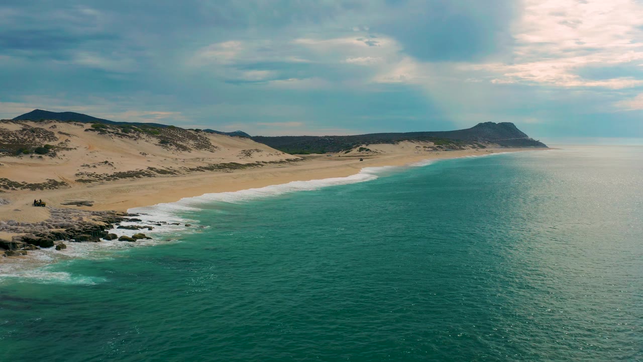 Aerial view at Laguna Beach with dark rocks at the lower left, teal water across the foreground, a pale sand beach through the center, inland dunes, and no visible people.