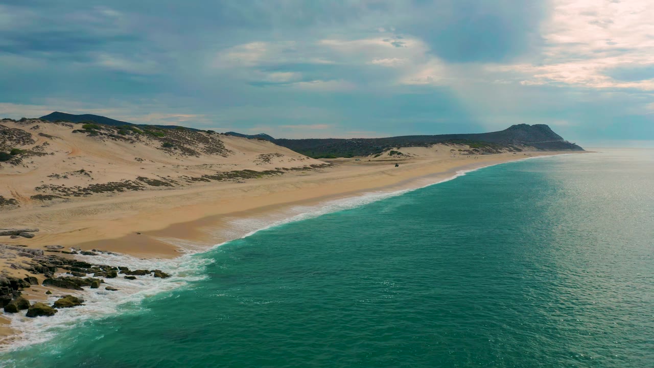 Aerial view at Laguna Beach, Baja California Sur, showing blue-green sea filling the right half, a narrow foam edge, pale sand, dune vegetation, distant hills, and no visible people.
