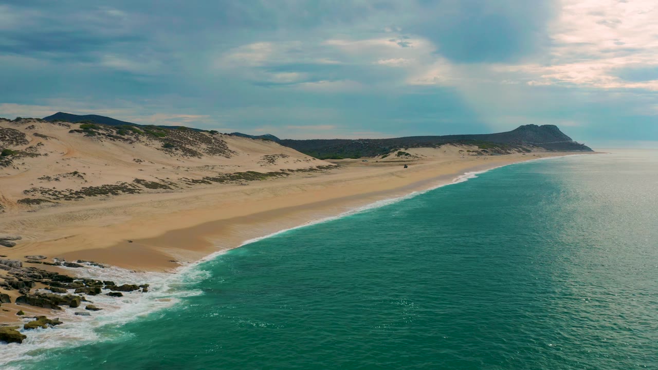 Drone-style aerial frame over Laguna Beach with the shoreline running diagonally, bright turquoise water, tan beach, scrub-covered dunes, a dark headland in the distance, and no visible people.
