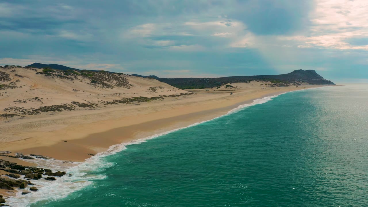 Aerial frame from Laguna Beach, Baja California Sur, where foamy waves spread over the lower shoreline, dunes rise inland, the sea stays calm to the right, and no visible people appear.