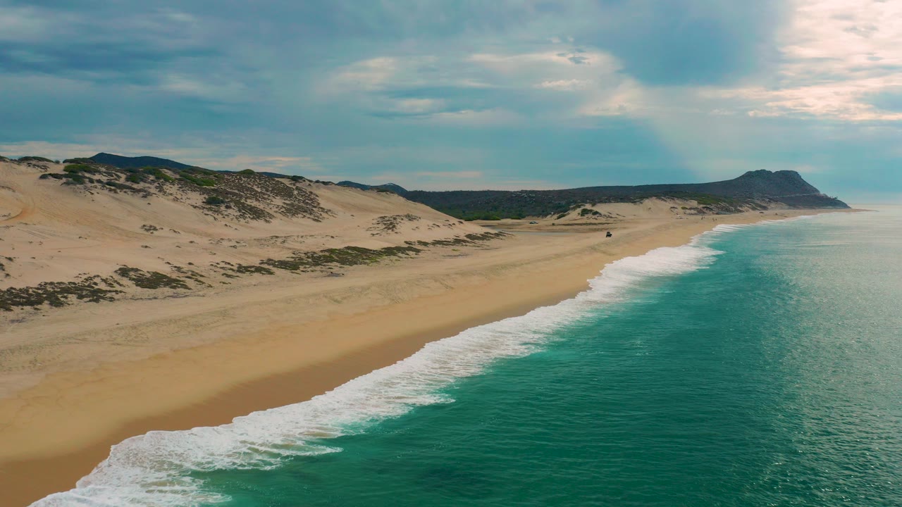 Opening aerial frame over Laguna Beach, Baja California Sur, with tan dunes at left, turquoise water at right, a distant headland, clouded sky, and no visible people.