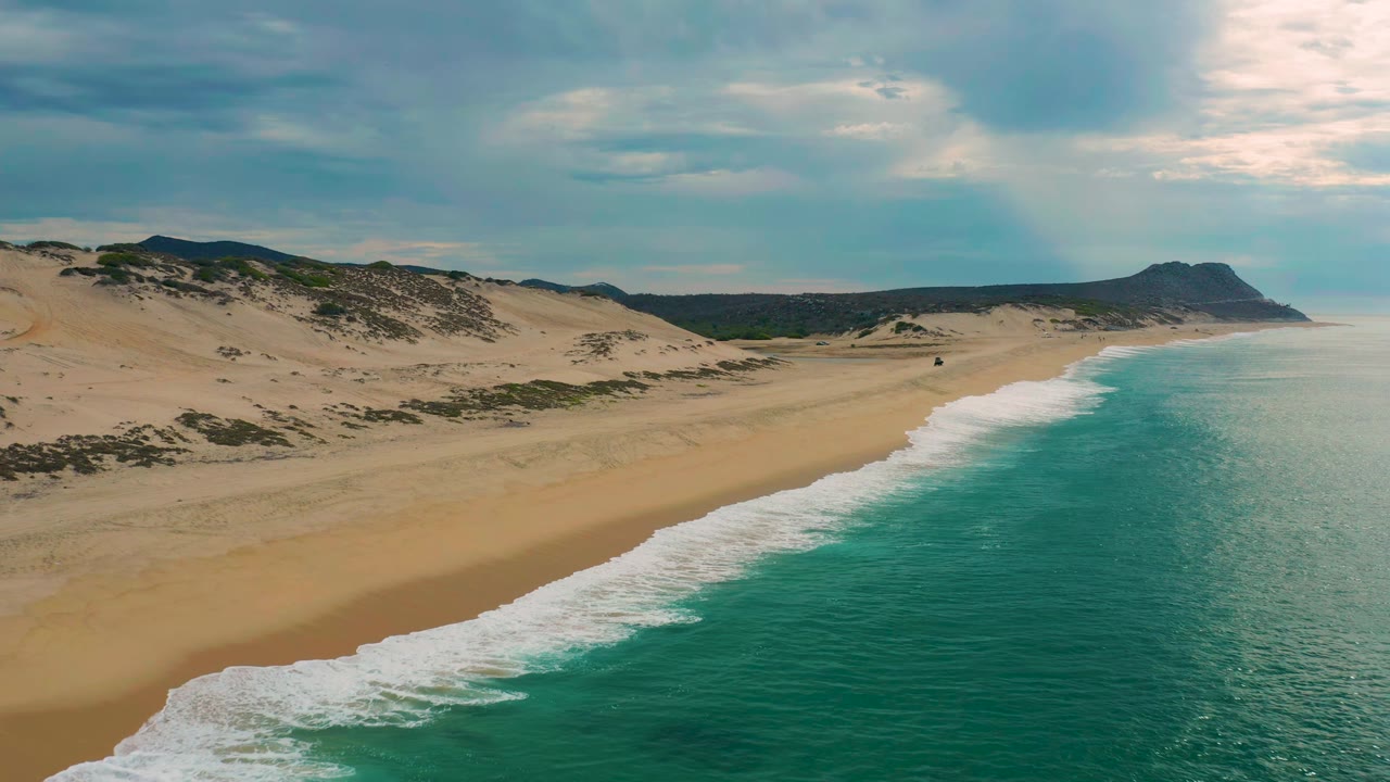 Aerial frame at Laguna Beach, Baja California Sur, showing the surf band closer to center, dune ridges inland, dark vegetation patches, distant headland, and no visible people.