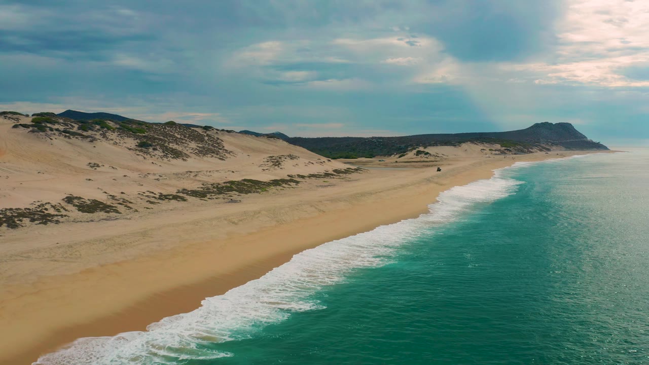 Aerial view of Laguna Beach, Baja California Sur, with a wide band of turquoise ocean, foamy shore break, pale sand, dune terrain, a distant headland, and no visible people.