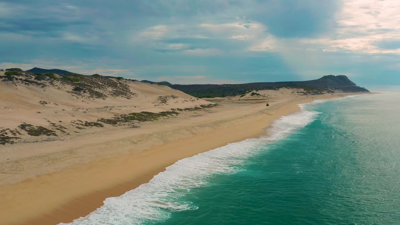 Mid-clip aerial frame over Laguna Beach, Baja California Sur, showing a longer stretch of empty beach, low dunes, green vegetation patches, pale surf, and no visible people.