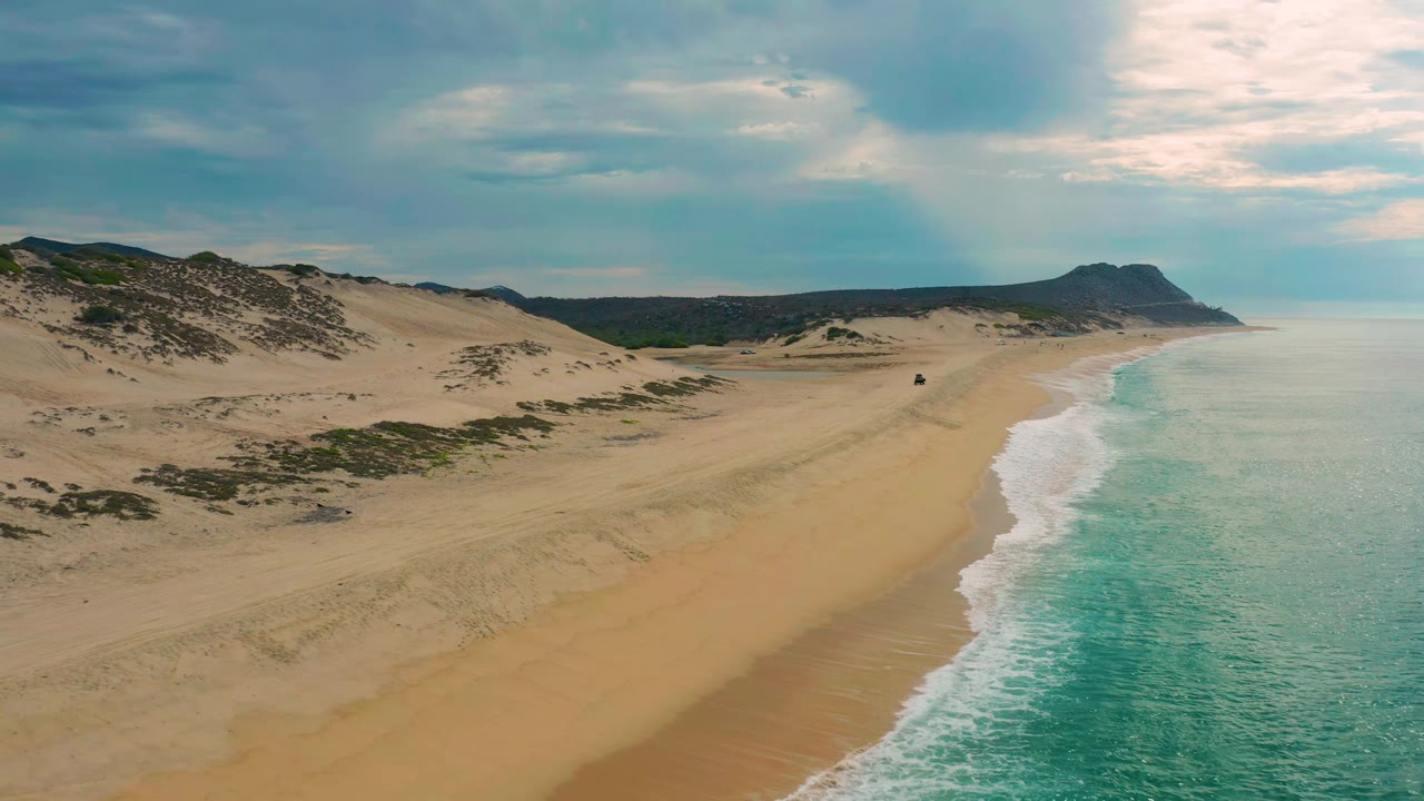 Aerial view of Laguna Beach, Baja California Sur, showing a long empty sand strip, a narrow white surf edge, turquoise water, low dunes, inland scrub, and no visible people.