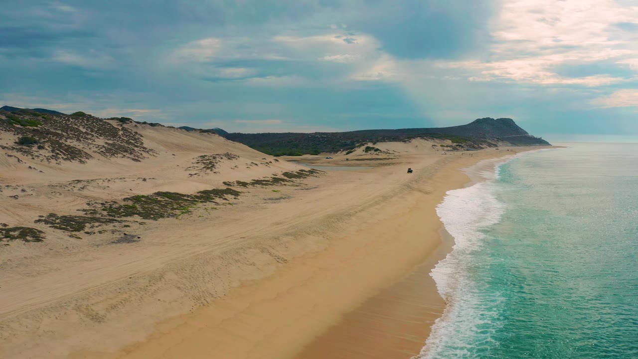Drone-height frame of Laguna Beach in Baja California Sur, with the sandy shoreline running into the distance, scrubby dunes on the left, pale turquoise water on the right, and no visible people.