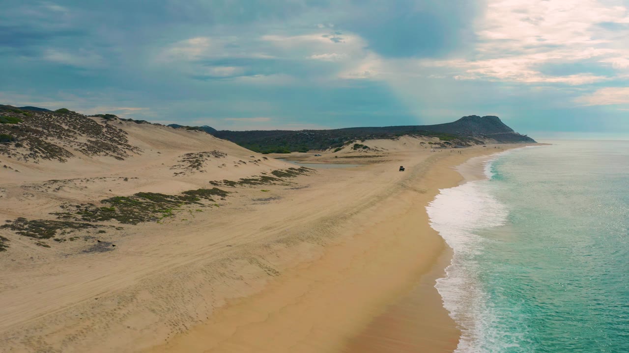 Closer aerial angle over dunes, empty sand, surf, and distant headland.