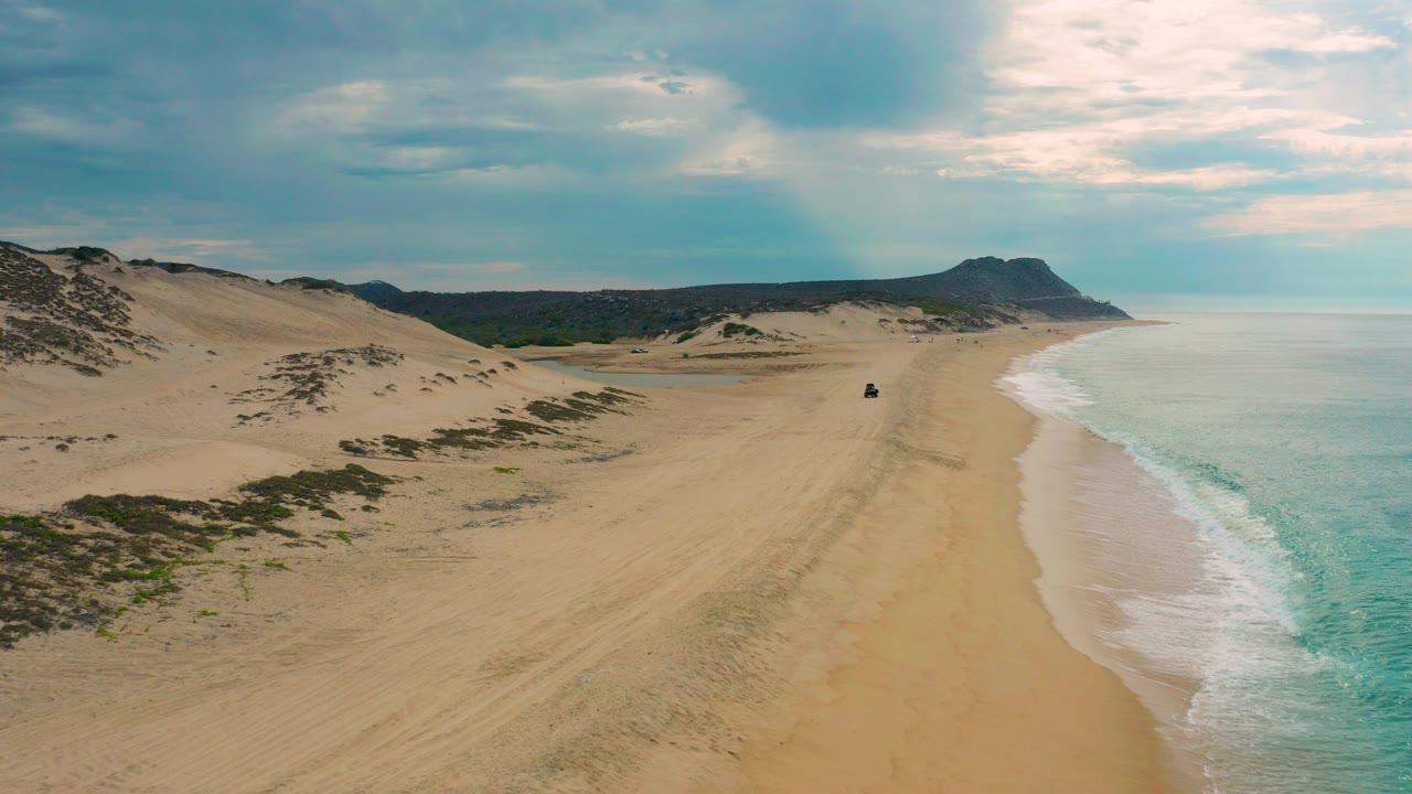 Aerial frame over Laguna Beach in Baja California Sur showing dunes at left, a wide sandy beach, blue-green surf at right, distant hills, cloudy light, and no visible people.