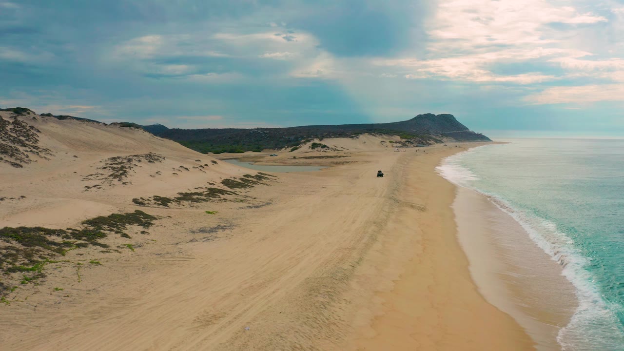 Aerial view over Laguna Beach in Baja California Sur showing pale sand stretching toward hills, foamy surf on the right, low dune vegetation on the left, cloud-filtered sky, and no visible people.