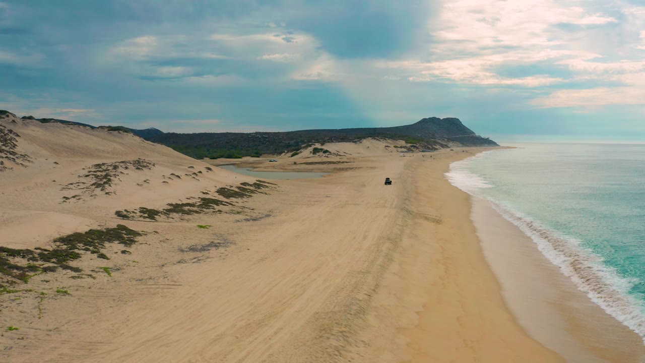 Mid-clip aerial frame of Laguna Beach, Baja California Sur with dune ridges at left, an open sandy track through the beach, surf washing the right side, a distant headland, and no visible people.