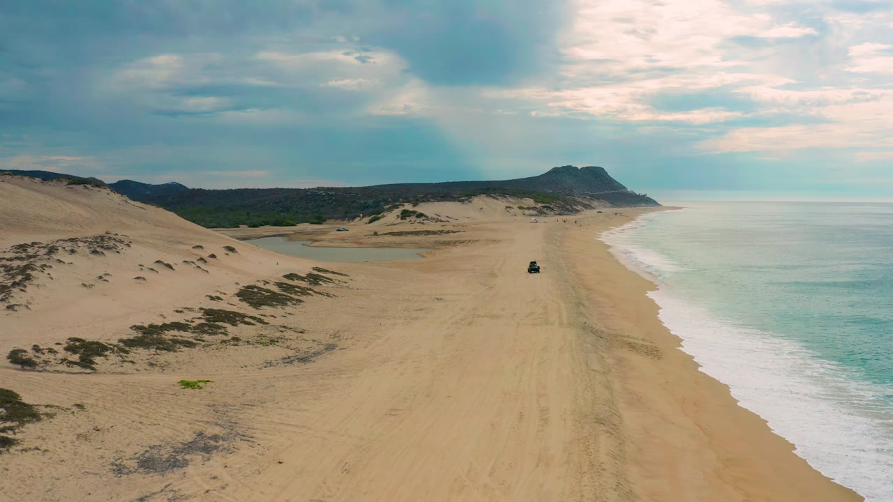 Aerial view over Laguna Beach, Baja California Sur, showing the beach stretching toward a low headland, a small dark vehicle on the sand, surf at right, and no visible people.