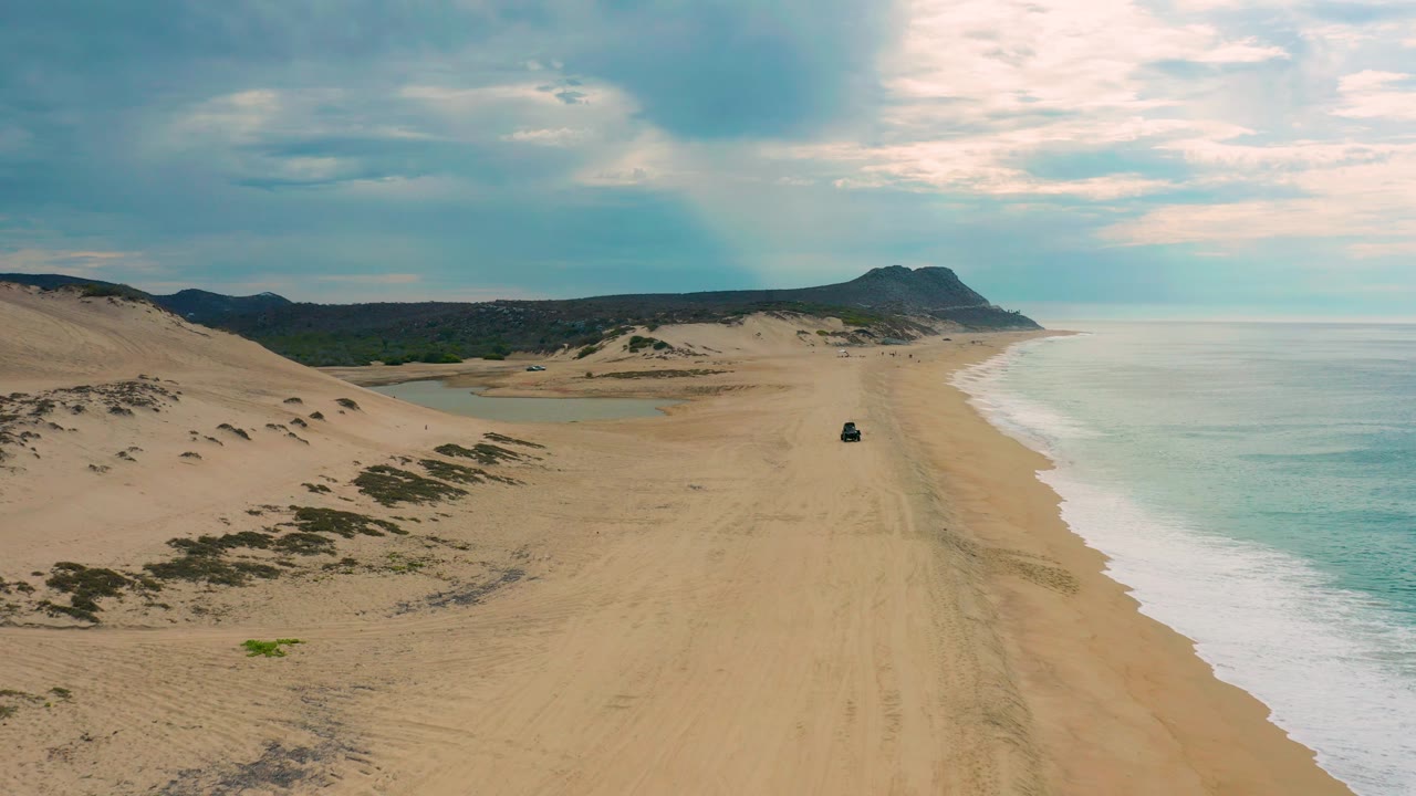 Wide aerial frame over Laguna Beach, Baja California Sur, with dune scrub at left, the surf line curving along the right edge, a dark vehicle on the beach, and no visible people.