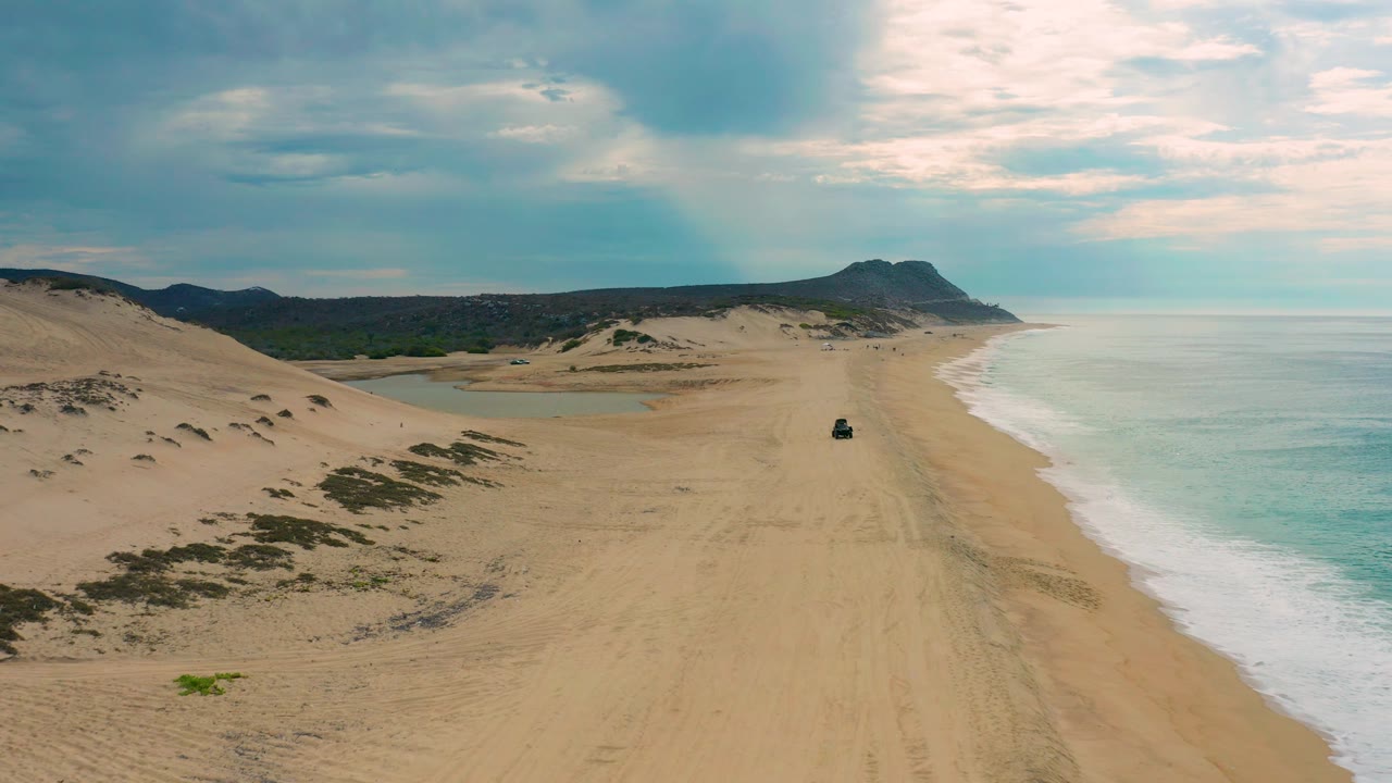 Drone frame over Laguna Beach, Baja California Sur, with a shallow inland water area near the dunes, broad sand, surf to the right, a distant mountain ridge, and no visible people.