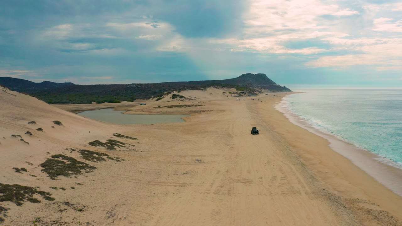 Opening aerial frame over Laguna Beach, Baja California Sur, showing sand dunes at left, a wide beach, pale surf at right, distant hills, and no visible people.